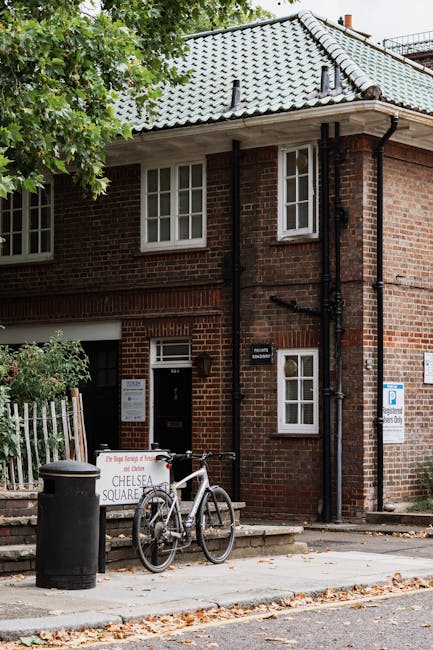 The image depicts a small, brick residential building with a grey-tiled, sloped roof and white-framed windows. In front of the house, there is a paved sidewalk lined with fallen leaves, indicating an outdoor urban setting. Leaning against a short stone step at the entrance, a white bicycle with black components is parked, next to a large black rubbish bin. The bin is positioned beside a white and red signboard that partially reads 'Chelsea Square,' suggesting a local area or street name, and appears to be related to waste or rubbish collection. To the left, part of a white wooden fence is visible, enclosing a small garden area with some green shrubbery. The facade of the building features black drainpipes running vertically down the wall, and the scene is shaded by leafy green trees overhead, creating a mix of natural and built environment. Overall, the setting effectively illustrates an example of private rubbish disposal in a residential context, aligning with the theme of alternative waste handling methods that Rubbish Removal Chelsea may support, especially in urban areas requiring independent collection solutions.
