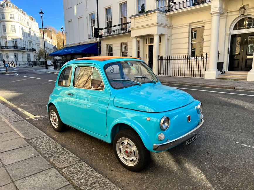 A vintage light blue compact car with a rounded shape, small size, and retro design is parked on the street in front of a white, multi-story building with black window frames and a black door marked with the number 18. The car has a beige fabric roof cover, chrome bumper, and hubcaps, with slightly tinted windows that reflect nearby structures. It is situated on a grey tarmac road with a textured concrete curb on the left, and the street is lined with black metal railings and traditional-style street lighting. In the background, there are other cars, pedestrians, and additional buildings with classic architectural details. The scene appears to be during daytime with clear, blue sky and natural sunlight illuminating the scene, creating subtle reflections on the car's glossy finish. This setting, combined with the presence of an vintage vehicle, subtly relates to private or alternative waste handling methods, as seen in the context of rubbish removal services provided by Rubbish Removal Chelsea at https://rubbishremovalchelsea.org, often involving on-site collections or specialized disposal arrangements.