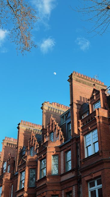 A clear daytime view of the upper section of a historic red brick building with ornate architectural details, including decorative battlements and arched windows. The building is situated on a street surrounded by bare tree branches extending into the frame from the top left corner. The sky is bright blue with some scattered white clouds, and the moon is visible in the distance slightly above and to the left of the building. The scene reflects an urban environment where private property and historic architecture are present, aligning with outside clearance or maintenance work that Rubbish Removal Chelsea might undertake in such settings. The overall atmosphere is calm with natural daylight illuminating the brickwork, highlighting its textures and finishes, suitable for visual context related to rubbish removal and on-site clearance services at Chelsea flats.