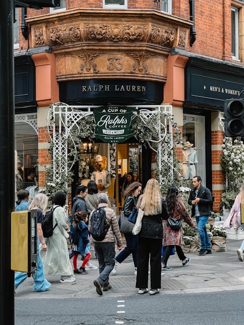 A group of pedestrians, including adults and children, are seen walking and standing outside a Ralph Lauren store on a busy city street. The store is situated in a red-brick building with an ornate stone facade above, featuring the number 35. The storefront is decorated with greenery and white flowers, and is framed by white decorative trellis panels. Inside the store, mannequins display clothing, and the interior lighting creates a warm glow. The scene suggests a vibrant retail environment, with people engaging in shopping or social activities, possibly related to private waste handling or rubbish removal events in commercial areas. The image highlights urban pedestrian activity in front of retail outlets, with visible details of clothing, accessories, and the surrounding architecture, aligning with the context of street-level commercial waste or rubbish management services involved in maintaining such environments. Rubbish Removal Chelsea might handle waste collection for similar retail locations in the area.