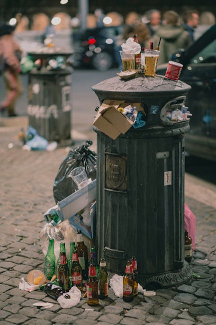 A black, cylindrical public trash bin with a domed lid is overflowing with discarded food containers, paper cups, and bottles, some placed on top and others spilling over the sides onto the cobblestone pavement. The waste includes plastic and paper waste such as crumpled paper, tissue, and a cardboard food container, with several glass bottles, some green and some brown, placed at the base of the bin. In the background, a blurred urban street scene is visible, with shoppers and vehicles, including a black car partially seen on the right side. A grey trash bag is tied and leaning against the waste bin, and additional litter is scattered around the base. The scene appears to be taken during daytime with natural lighting, highlighting the mess overflowing from the bin, which highlights the importance of efficient rubbish removal services like those provided by Rubbish Removal Chelsea, especially for managing waste in busy public areas while supporting alternative waste handling solutions.