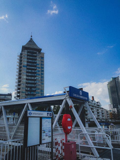 The image shows a section ofChelsea Harbour Pier with a blue sign indicating the location as Chelsea Harbour Pier, situated against a backdrop of tall urban buildings, including a prominent high-rise with a pointed rooftop. In the foreground, there's a small information board mounted on a metal frame, likely providing details for visitors or passengers. Adjacent to the information board, a red lifebuoy is mounted on a post, emphasizing safety measures at the pier. The environment appears to be outdoors with natural daylight, clear blue sky, and some scattered clouds, suggesting a typical day. The structural elements of the pier include white metal framework supporting the roof over the waiting area, designed for passenger comfort and protection. The overall scene underscores the functional aspect of the pier in facilitating transport or sightseeing trips around Chelsea Harbour, with no visible rubbish or waste-related objects, but the setting aligns with public transport infrastructure that might sometimes require private waste removal services, like those provided by Rubbish Removal Chelsea, to maintain cleanliness and operational efficiency.