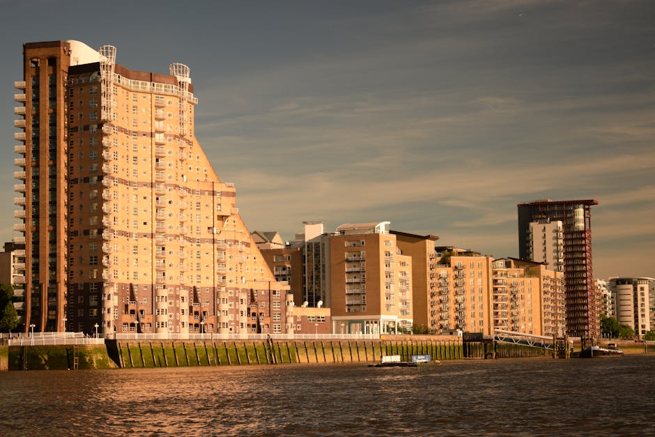 The image shows a section ofChelsea Harbour Pier with a blue sign indicating the location as Chelsea Harbour Pier, situated against a backdrop of tall urban buildings, including a prominent high-rise with a pointed rooftop. In the foreground, there's a small information board mounted on a metal frame, likely providing details for visitors or passengers. Adjacent to the information board, a red lifebuoy is mounted on a post, emphasizing safety measures at the pier. The environment appears to be outdoors with natural daylight, clear blue sky, and some scattered clouds, suggesting a typical day. The structural elements of the pier include white metal framework supporting the roof over the waiting area, designed for passenger comfort and protection. The overall scene underscores the functional aspect of the pier in facilitating transport or sightseeing trips around Chelsea Harbour, with no visible rubbish or waste-related objects, but the setting aligns with public transport infrastructure that might sometimes require private waste removal services, like those provided by Rubbish Removal Chelsea, to maintain cleanliness and operational efficiency.
