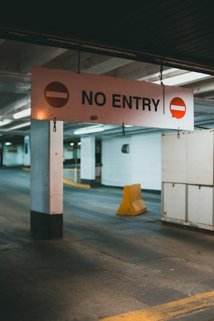 A parking garage interior featuring a large rectangular sign hanging from the ceiling that reads 'NO ENTRY' in bold black letters, accompanied by two circular symbols: on the left, a black circle with a horizontal white line indicating no access, and on the right, a red circle with a white horizontal bar. The sign is mounted on metal hooks attached to a support beam beneath a dark, ribbed ceiling. In the background, the garage's concrete walls and floor are visible, with painted yellow lines marking parking spaces and drive lanes. A concrete pillar painted white with a black base stands to the left of the sign. On the right, a white barrier with a metal railing section is partially visible. The overall environment is dimly lit, with artificial lighting casting soft reflections on the concrete surfaces. This scene exemplifies a restricted area within a private parking structure, relevant to waste management services managing access to rubbish removal or disposal areas that require adherence to access limitations, aligned with the context of alternative waste handling solutions.