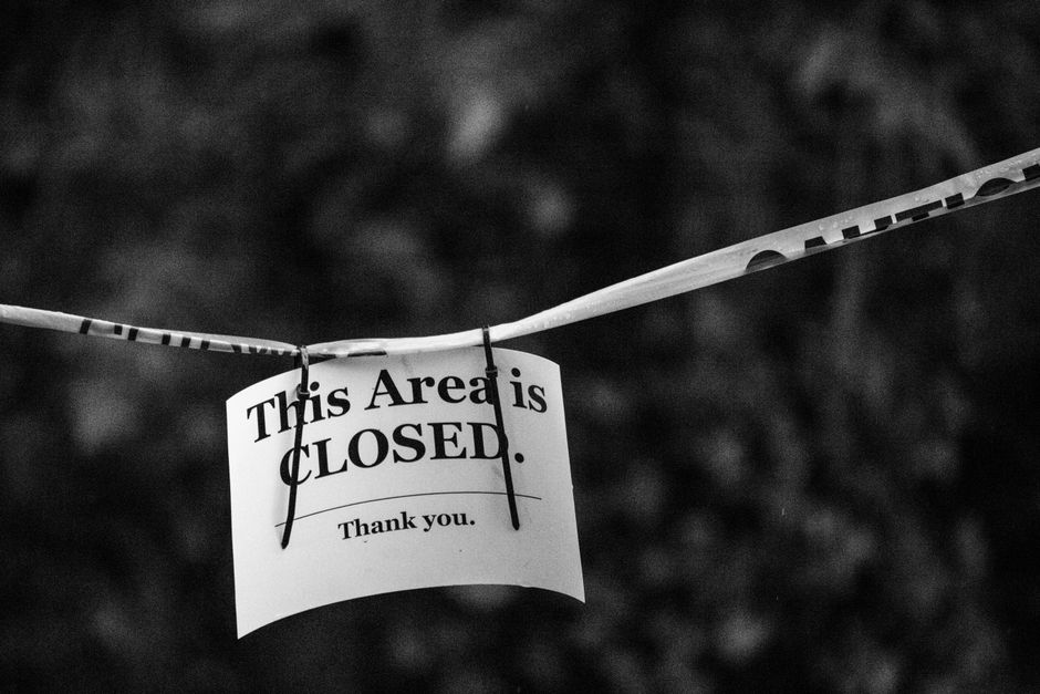 A close-up black and white photograph showing a sign attached to a tape or string across a natural outdoor setting, with text stating 'This Area is CLOSED. Thank you.' The sign appears to be made of paper or lightweight material, mounted with two small black clips or pins at the top corners. The background is blurred, suggesting trees or foliage, indicating an outdoor environment. The tape or string is stretched horizontally and is partially loose or twisted, emphasizing the restriction or closure of the area, which may relate to site safety or restricted access in waste management or clearance contexts. The lighting is diffuse, with no harsh shadows, contributing to a neutral and professional tone suitable for accessibility and service-related descriptions. This scene subtly underscores the importance of controlled entry or restrictions, which are relevant considerations for private rubbish removal or on-site clearance services provided by Rubbish Removal Chelsea.
